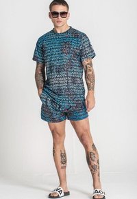 Krátký rukáv a šortky v soupravě s vícebarevným grafickým vzorem písmen. Materiál vypadá lehký; nošeno s nazouvacími sandály.