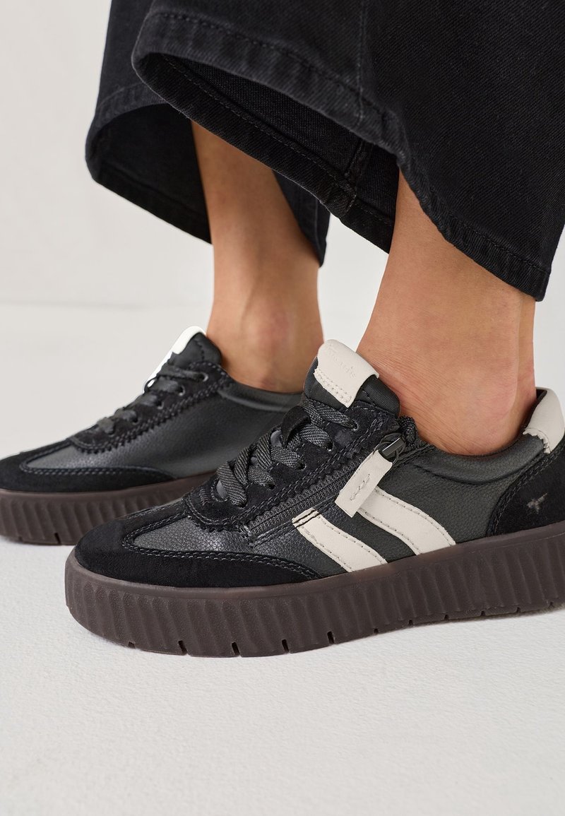 Baskets basses noir et blanc avec fermeture éclair latérale, portées avec un pantalon court noir, sur une surface plate blanche.