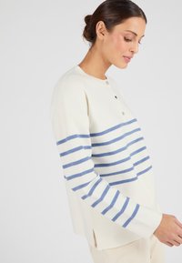 Witte trui met marineblauwe strepen, ronde halslijn, drieknoopsluiting en een ontspannen pasvorm. Zachte textuur met zijsplitten aan de zoom.