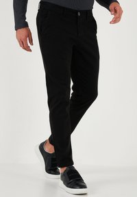 Pantaloni negri dintr-un material elastic, cu o croială slim, având buzunare laterale și închidere cu nasture, asortați cu teniși negri și o cămașă gri cu mâneci lungi.
