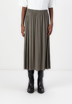 Femme portant une jupe midi plissée taille haute vert olive associée à des bottes noires brillantes hauteur genou et un haut blanc rentré.