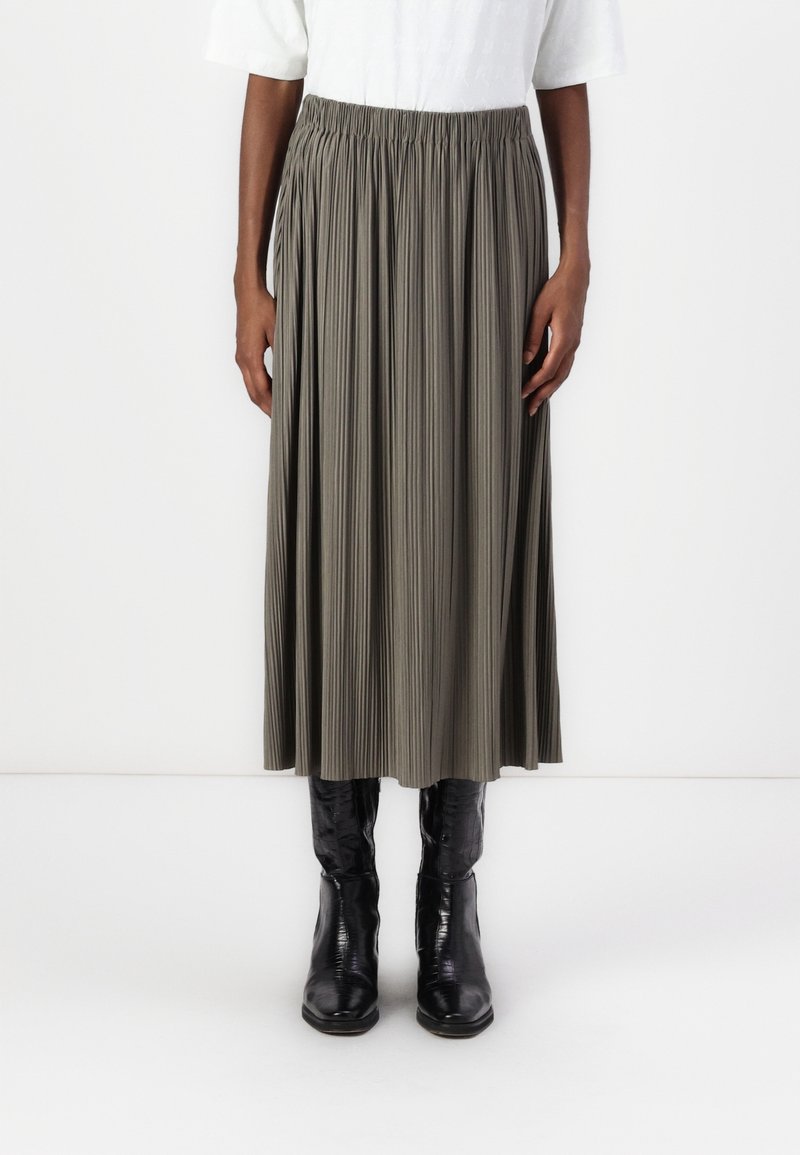 Femme portant une jupe midi plissée taille haute vert olive associée à des bottes noires brillantes hauteur genou et un haut blanc rentré.