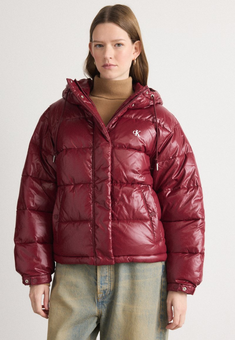 Burgunderfarget pufferjakke med en skinnende finish, hette, frontglidelås, justerbare snorer og logo. Brukt over en lys topp og lyse denimbukser.