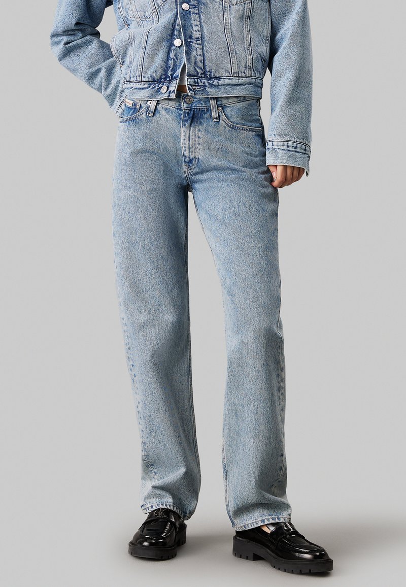 Jeans de mezclilla azul claro con un corte de pierna recta, costuras visibles, diseño de cinco bolsillos y textura desgastada, combinados con zapatos de plataforma negros brillantes.