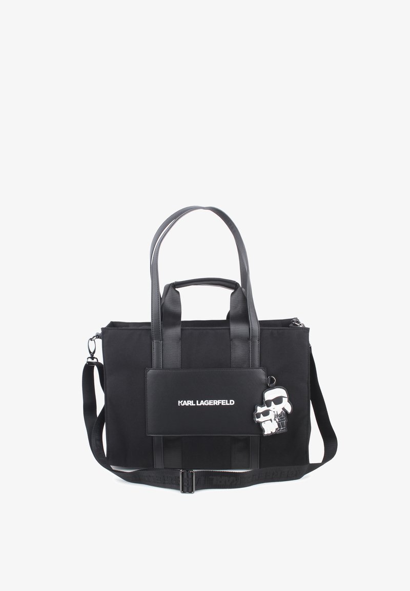 Black tote bag featuring fabric material, leather accents, two top handles, and an adjustable shoulder strap. Includes a logo patch and charm detail.