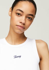 Top de tirantes blanco con canalé que presenta un escote redondo y un logotipo bordado en navy de "Tommy" en el pecho. Textura suave, diseño ajustado.