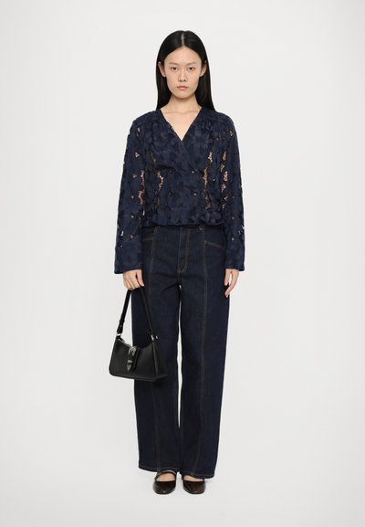 Navy lace blouse with flower pattern, cropped length, long sleeves; paired with high-waisted dark denim pants and a small black handbag.