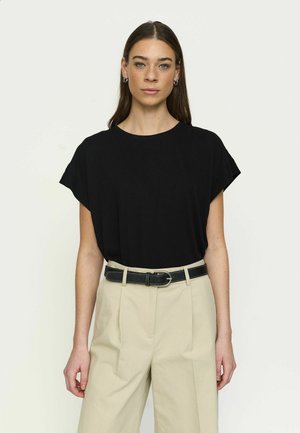 Kobieta o prostych brązowych włosach ubrana w czarny krótki top z rękawami, włożony do beżowych spodni z wysokim stanem i czarnym paskiem, stoi na jednolitym tle.