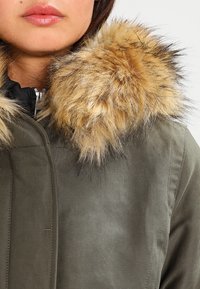 Chaqueta verde oliva con un cuello de piel sintética desmontable en tonos marrón claro y oscuro, que cuenta con cierre de cremallera y textura acolchada.