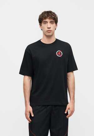 Giovane uomo con capelli castani ricci che indossa una maglietta nera con un piccolo logo circolare rosso e bianco sul petto sinistro.