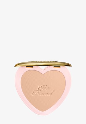 Heart-shaped compact powder with "Too Faced" embossed on beige makeup, pink case, and gold hinged lid.