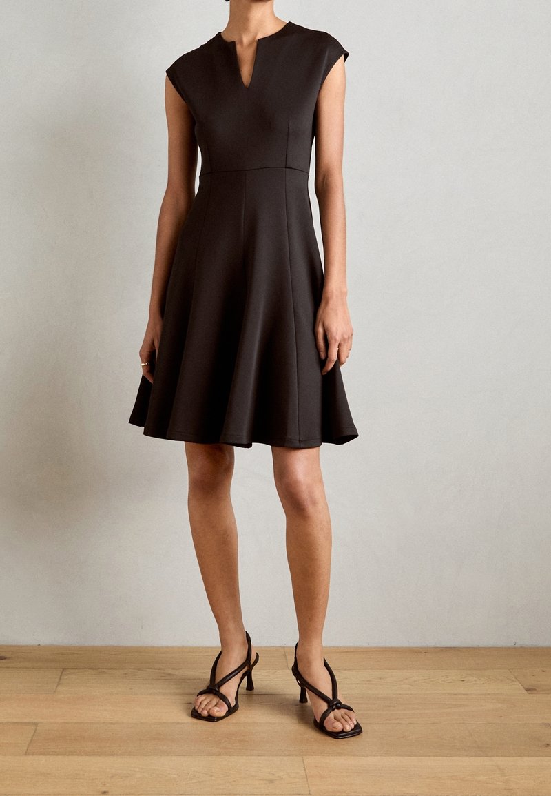 Robe noire avec un corsage ajusté, des manches courtes et une jupe évasée. Elle présente un décolleté en œillet et un tissu lisse et texturé.