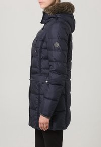 Giubbotto puffer blu navy con cappuccio foderato di pelliccia, polsini a costine, cuciture verticali e dettagli con bottoni a pressione. Presenta un logo sulla manica sinistra.