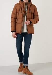 Brun pufferjacka med huva, med två främre dragkedjor, elastiska ärmändar och en quiltad design. Bärs över en ljus tröja och blå jeans.