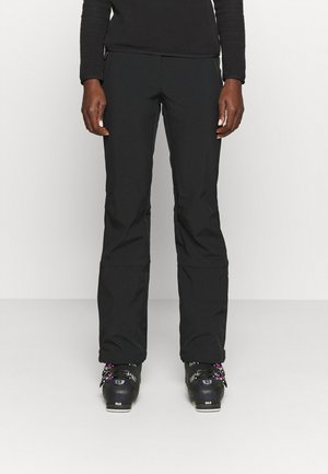 Personne portant un pantalon de ski noir et des chaussures de ski noires, debout devant un fond clair uni.