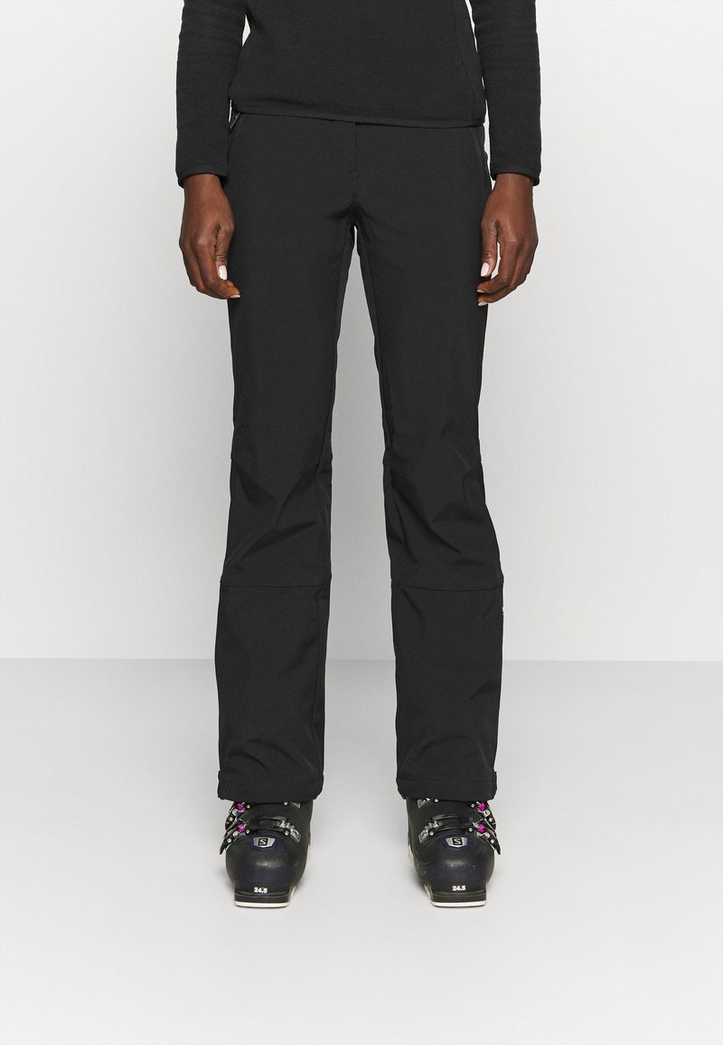 Personne portant un pantalon de ski noir et des chaussures de ski noires, debout devant un fond clair uni.