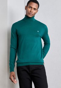 Pull à col roulé turquoise en tissu doux, avec des manches longues et un ourlet à côtes, associé à un pantalon noir. Petit logo sur la poitrine.
