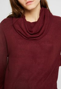 Haut bordeaux à col châle en tissu doux, avec un col drapé et des manches longues, offrant une texture lisse et uniforme.