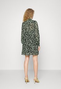 Vapauttava musta mekko, jossa on vaaleanvihreä abstrakti kuvio, pitkät hihat, napitettu hihansuu ja suora helma, yhdistettynä vaaleanvihreisiin korkokenkiin.