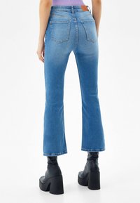 Světle modré zvonové džíny vyrobené z denimu, s středním pasem, pěti kapsami a hnědou koženou nášivkou na zadním pásu.