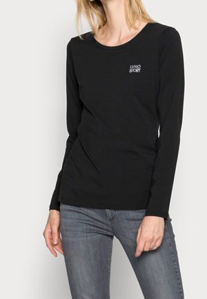 Femme portant un t-shirt noir à manches longues avec le texte "UNO SPORT" sur la poitrine et un jean gris ajusté, debout devant un fond blanc.