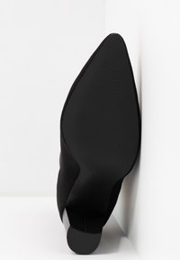 Bottine noire à bout pointu avec empeigne en tissu côtelé et petit talon bloc, positionnée semelle vers le haut contre des murs blancs d'angle.