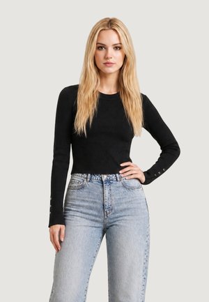 Donna con capelli biondi lunghi che indossa un top nero aderente a maniche lunghe e jeans azzurri a vita alta, in piedi con una mano sul fianco.