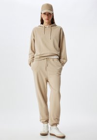 Beige casual outfit bestaande uit een hoodie en sweatpants. De hoodie heeft een trekkoord, en de broek heeft een elastische tailleband en boorden. Witte sneakers worden gedragen.