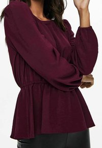 Burgundy blus med rund halsringning, elastisk midja och långa puffärmar. Tillverkad av texturerat tyg, har den en avslappnad passform.