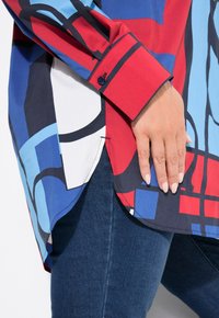 Blusa a maniche lunghe in rosso, blu e bianco con motivi astratti. Presenta un orlo curvo e polsini con bottone, realizzata in tessuto leggero.