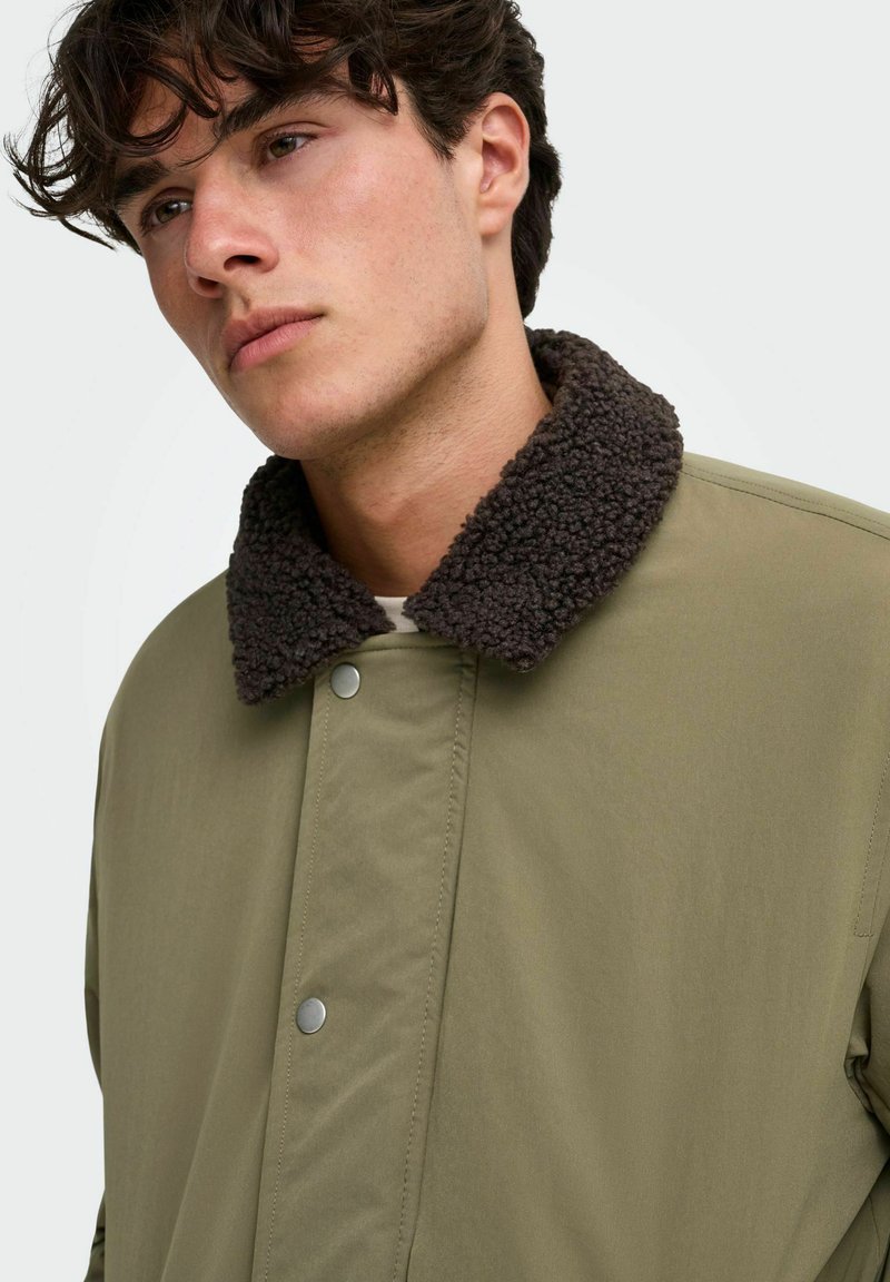 Jeune homme aux cheveux bouclés foncés portant une veste vert olive avec un col texturé foncé, regardant légèrement vers la gauche sur un fond blanc.