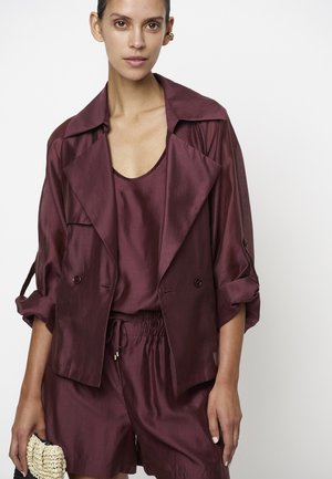 Vrouw met een bordeauxrode satijnen jas, bijpassende losse korte broek en top, houdt een zwarte en beige geweven clutch vast tegen een witte achtergrond.