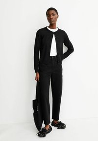 Cardigan noir à col rond avec des boutons, porté sur un t-shirt blanc. Associé à un pantalon noir taille haute et des chaussures à franges.