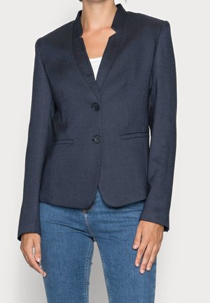 Femme portant un blazer bleu marine foncé ajusté avec un col cranté et deux boutons sur un top blanc et un jean bleu.