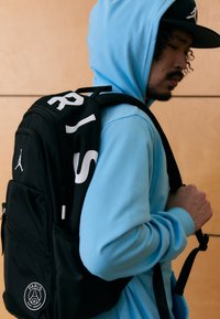 Fekete hátizsák fehér "PARIS" felirattal és egy logóval elöl. Simább anyagból, cipzáras zsebekkel, állítható pántokkal, egy kék kapucnis felsőt viselő személy által hordva.