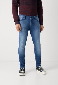 Jeans de denim azul ajustados con textura descolorida, diseño de cinco bolsillos y costuras en contraste, combinados con zapatillas de lona negras.