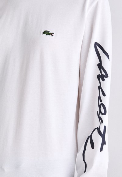 T-shirt à manches longues blanche, arborant le logo crocodile brodé en vert et un accent en texte en cursive noire sur la manche. Texture lisse en coton.