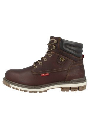 Botas de tobillo de cuero marrón con suelas de goma marrón, ojales de metal y cordones. Incluyen un collar acolchado y una etiqueta de logo roja.