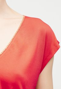 Κοραλλί κοντομάνικη μπλούζα με λαιμόκοψη σε σχήμα V, διακοσμημένη με μικρές μπεζ χάντρες, φοράει ένα άτομο με ανοιχτόχρωμο δέρμα.