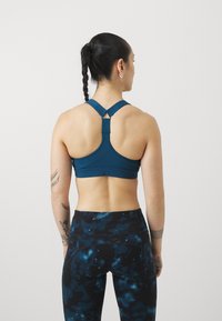 Tirkīza sporta krūšturis ar racerback dizainu un regulējamām siksnām, apvienots ar melniem legingiem, kuriem ir zils abstrakts raksts.