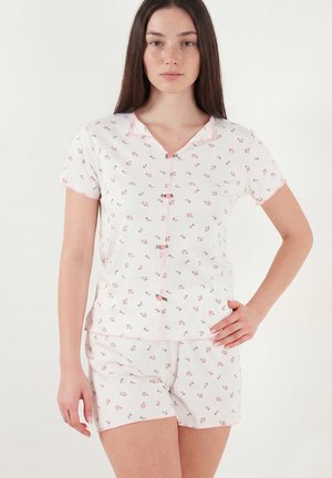 Young woman wearing white short-sleeve buttoned top and matching shorts with small pink floral pattern, standing with one hand on hip.