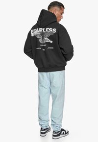 Schwarzer Kapuzenpullover mit großen weißen "FEARLESS"-Schriftzug und einem Adler-Design auf dem Rücken, kombiniert mit hellblauen Jeans und schwarz-weißen Sneakers.