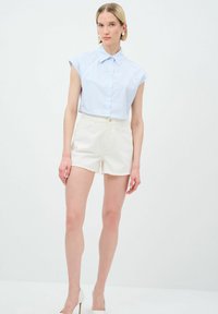 Camicia azzurra a maniche corte con bottoni e colletto, abbinata a pantaloni corti in denim bianchi con orlo sfrangiato. I tacchi a spillo completano il look.