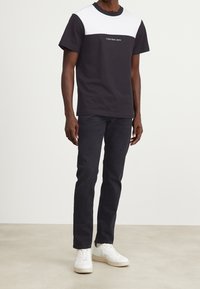 Camiseta de bloques de color en blanco y negro con un cuello redondo y un logotipo en el frontal. Combinada con jeans oscuros y zapatillas blancas.