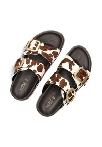 Koeienprint slide sandalen met een bovenkant van bruin en wit faux bont, zwarte rubberen zool, enkele gesp-sluitingen en goudkleurige hardware.