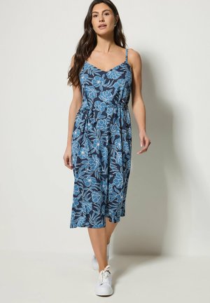 Vrouw die loopt en een blauwe midi-jurk met bloemenprint, dunne bandjes en witte sneakers draagt tegen een effen lichte achtergrond.