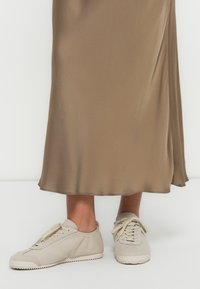 Μπεζ αθλητικά παπούτσια με κορδόνια, ανώτερο μέρος από σουέτ με υφή και καουτσούκ σόλα, συνδυασμένα με μια μακριά, ρέουσα φούστα από σατέν σε ελιά πράσινο χρώμα.