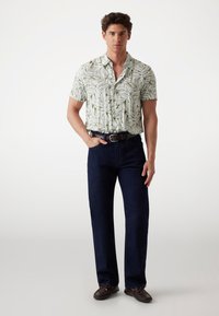 Camisa de manga corta con estampado de hojas verdes sobre fondo blanco, combinada con pantalones azul oscuro y zapatos mocasines marrones. Ajuste casual.