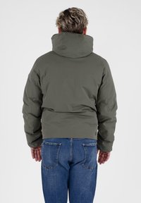 Groene gewatteerde jas met een hoge kraag, gladde textuur en een verkorte snit. Draagt om met blauwe jeans, waarop de pocketdetails aan de achterkant zichtbaar zijn.