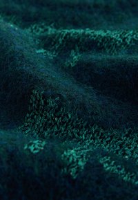 Teksturirana tkanina v globokih odtenkih modro-zelene in modre barve, ki prikazuje različne vzorce pletenja z mehkim, puhastim videzom.